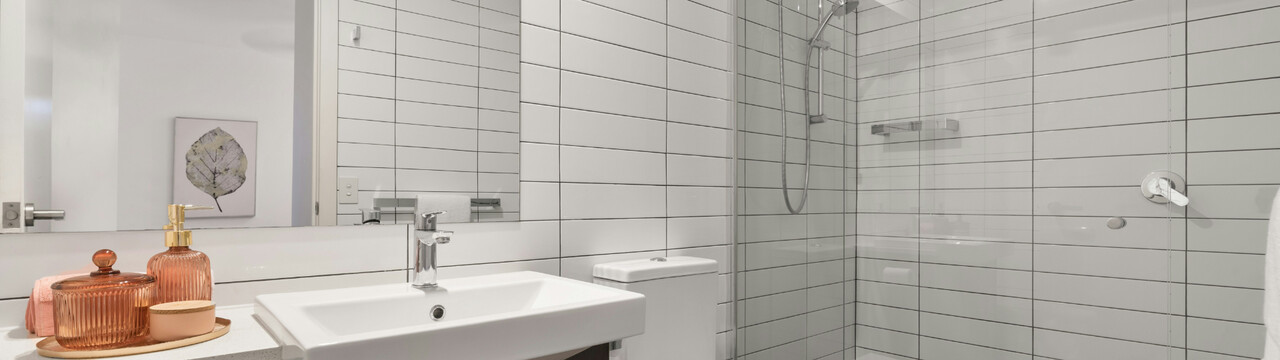 Blick in ein Badezimmer mit weiß gefliesten Wänden. Auf der rechten Seite im Bild sieht man eine bodentiefe Dusche, welche mit einer Glaswand vom Raum getrennt wird. Daneben ist eine Toilette erkennbar, welche zwischen Dusche und Waschbecken an der Wand ist. Über dem weißen, eckigen Waschbecken hängt ein großer Spiegel, in welchem sich eine Handtuchstange und die offene Badezimmer-Türe spiegelt. Durch die offene Tür sieht man in einen weißen Flur, an dessen Wand das Bild eines Blattes in schwarz-weiß hängt.