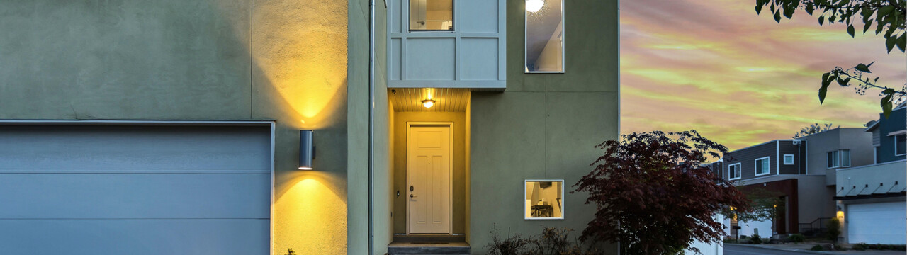 Detailaufnahme einer Außenwand, welche grün verputzt ist. Links im Bild sieht man in Nahaufnahme einen Teil der Fassade mit Garage. Auf der Fassade ist rechts neben dem Garagentor eine Lampe angebracht, welche nach oben und unten strahlt. Neben der Lampe ist ein Versatz des Hauses nach hinten sichtbar, sodass man aus mehr Entfernung die weiße Eingangstür und zwei Fenster sieht. Auf der rechten Seite des Bildes sieht man in eine Straße neben dem Haus, auf der auf der anderen Straßenseite weitere Häuser sind