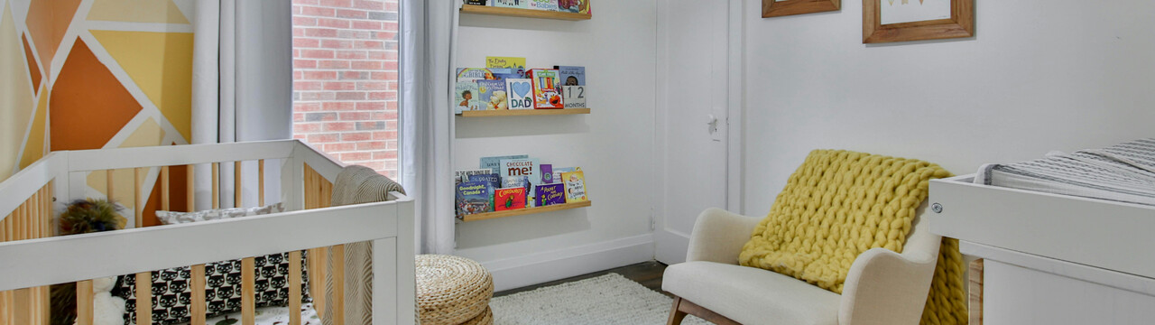 Blick in ein Kinderzimmer, in dem auf der linken Seite ein Babybett steht. Links erkennt man vorne eine Wickelkommode und daneben steht an der Wand ein halbhoher Schaukelstuhl. Über dem Schaukelstuhl an der Wand hängen Bilder. Neben der Tür hängen Regalbretter mit Büchern darauf und daneben ist ein Fenster mit Vorhängen. An der Wand über dem Babybett ist die Wand abstrakt mit verschiedenen Rot-, Orange- und Gelb-Tönen gestaltet. Auf dem Boden liegt ein heller Teppich.