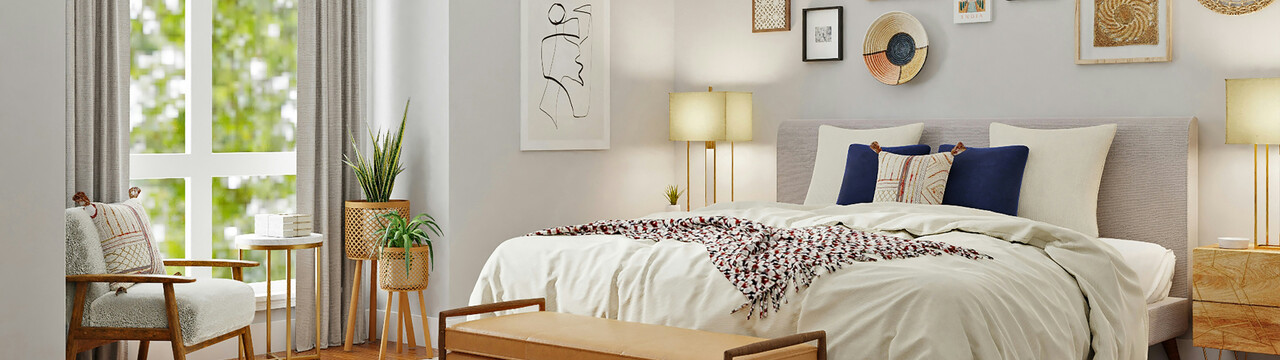 Blick in ein Schlafzimmer mit großem Bett, auf dem eine Decke und verschiedene Kissen liegen. Über dem Bett hängt verschiedene Deko an der Wand. Neben dem Bett rechts steht ein Nachttisch, auf dem eine Lampe leuchtet. Auf der linken Seite des Bettes sieht man eine identische Lampe. Links im Bild ist ein bodentiefes Fenster mit grauen Vorhängen, vor dem ein Sessel und ein Beistelltisch stehen. Neben dem Beistelltisch an der Wand stehen noch zwei Pflanzen.