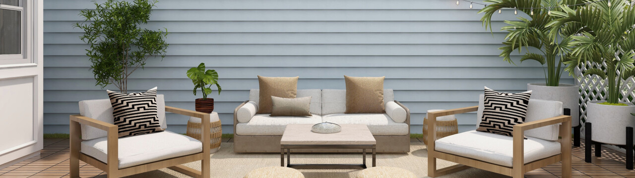 Blick auf eine Terrasse, in deren Hintergrund ein hellblauer Lattenzaun bis zum oberen Bildrand zu sehen ist. Davor steht ein Gartensofa mit hellen Polstern und links und rechts sind vier verschieden Pflanzen in Töpfen verteilt. Vor dem Sofa steht ein Gartentisch auf einem Außenteppich und im Vordergrund stehen zwei Sessel mit hellen Polstern, welche links und rechts schräg Richtung Kamera gedreht sind. Um den Teppich herum sind Terrassenfliesen zu erkennen und am linken Bildrand ist eine Häuserecke.