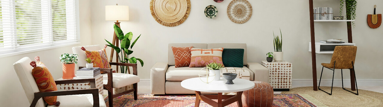 Blick in ein Wohnzimmer mit hellen Wänden, großen Fenstern auf der linken Seite des Bildes. Vor dem Fenster stehen zwei Sessel mit Holzgestell und hellen Polstern. Mittig im Bild ist ein helles Sofa zu sehen mit bunten Kissen darauf und davor ein runder Couchtisch mit Holzfüßen und ein Sitzkissen auf dem Boden. Über dem Sofa hängt runde Dekoration an der Wand und rechts neben dem Sofa steht ein heller Block mit zwei Pflanzen drauf. Links neben dem Sofa steht eine Lampe und davor eine größere Pflanze.