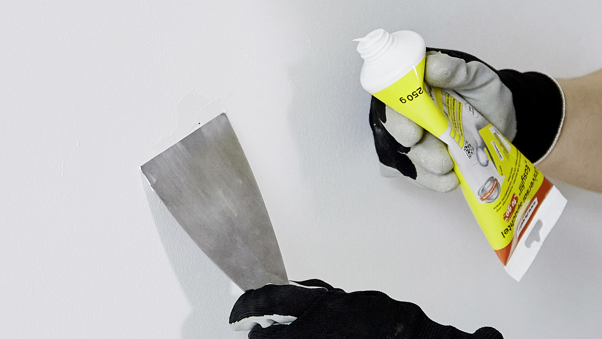 Nahaufnahme auf eine Hand mit Spachtel, welche an Spachtel an der Wand glatt zieht. In der anderen Hand ist eine Tube des Weber Easyfills sichtbar