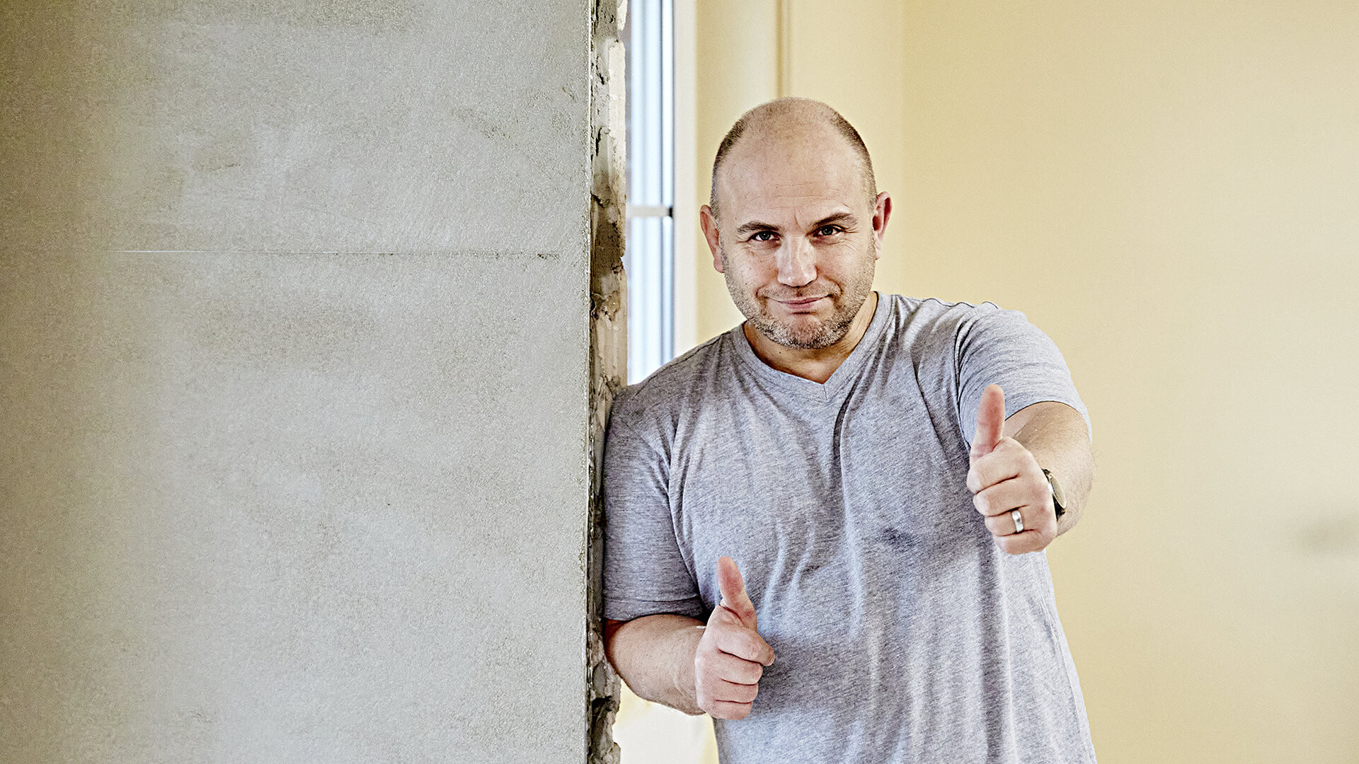 Mann lehnt an eine fertig verputzte Wand an und hält beide Daumen hoch in die Kamera
