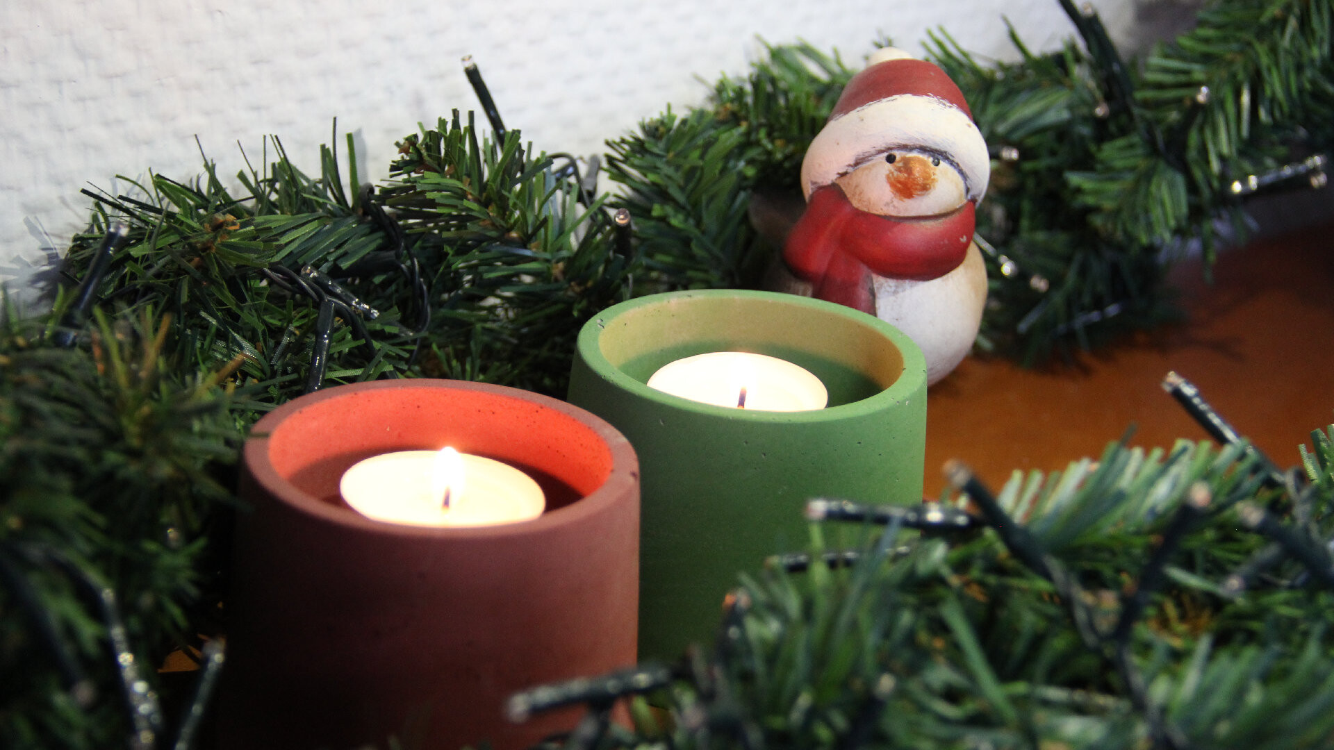 Aufnahme von Kerzenhaltern aus gefärbtem Bastelbeton in rot und grün mit Teelichtern drin. Die Kerzenhalter stehen auf einem Holztisch. Um diese herum liegt eine Lichterkette mit unechter Tannenzweig-Optik und hinter den Kerzenhaltern steht ein kleiner Schneemann aus Porzellan.
