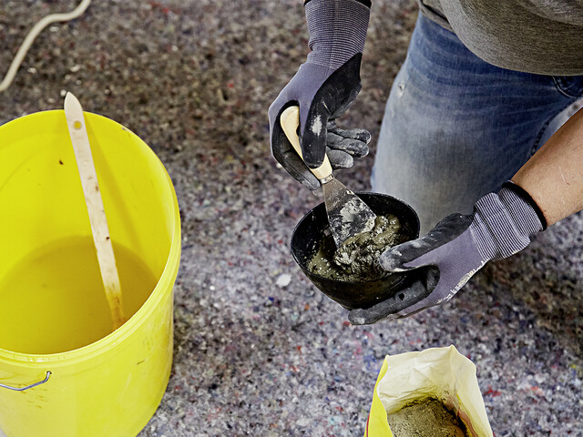 Nahaufnahme darauf, wie der Blitzzement in einem Gipsbecher angerührt wird. Dazu sieht man in der Mitte des Bildes zwei Hände, die mit dem Spachtel den Blitzzement in dem Gipsbecher verrührt. Unten links im Bild sieht man einen gelben Eimer mit Wasser und dem Flachpinsel drin. Im Hintergrund sieht man eine Fließunterlage.