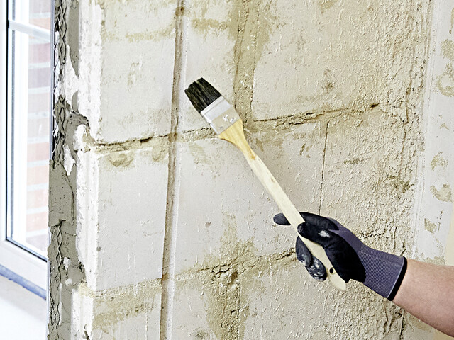 Nahaufnahme, wie der Leitungsschlitz vorgenässt wird. Dazu sieht man die Wand mit dem Leitungsschlitz im Hintergrund und davor eine Hand, welche einen Flachpinsel hält, welcher in die Richtung des Leitungsschlitz ausgerichtet ist
