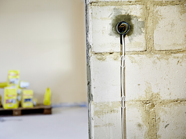 Aufnahme der Wand mit Unterputzdose und Leitungsschlitz mit Kabel im Vordergrund auf der rechten Seite. Auf der linken Seite sind im Hintergrund verschiedene Produktverpackungen sichtbar