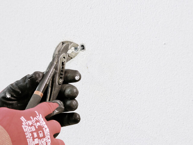 Nahaufnahme, wie eine Hand in Arbeitshandschuhen mit einer Zange eine Schraube greift, die in einen Dübel im Bohrloch in der Wand gedreht ist