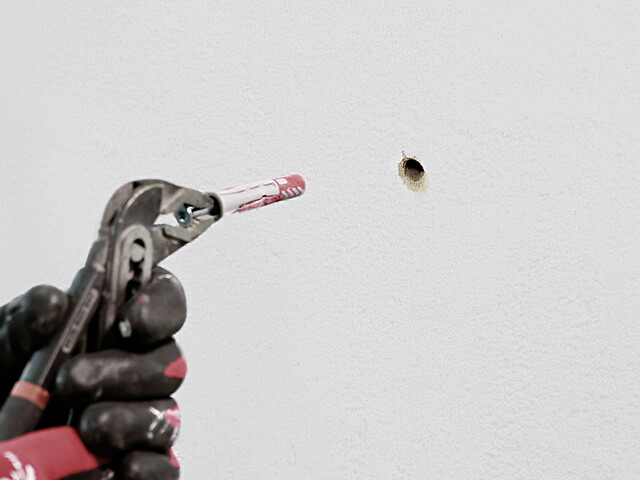 Nahaufname, wie Hand in Handschuhen mit einer Zange einen Dübel mit Schraube vor einem Bohrloch in der Wand hält
