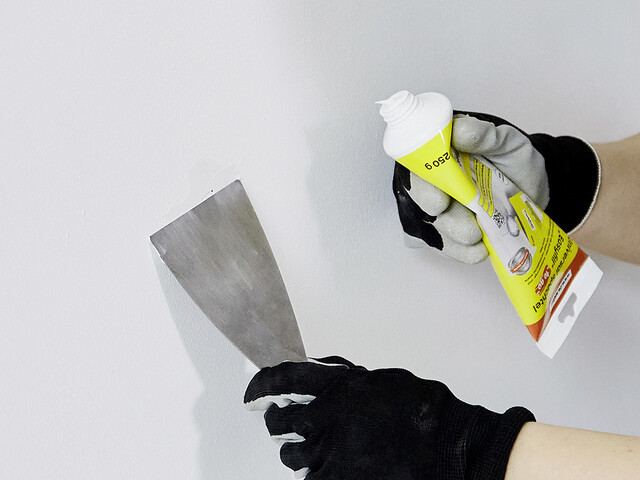 Nahaufnahme, wie eine Hand in Arbeitshandschuhen mit einem Spachtel über ein Loch in der Wand geht. In der anderen Hand ist eine Tube des Universal Spachtel Easyfill "to go" sichtbar