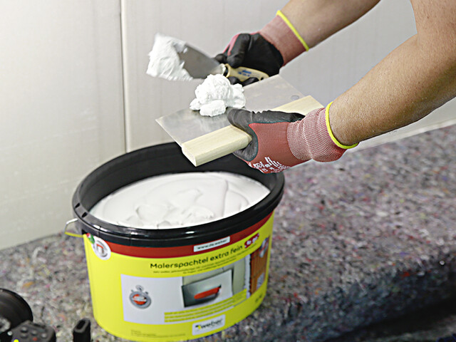 Nahaufnahme, wie der gebrauchsfertige Malerspachtel aus dem Eimer entnommen wird. Dazu steht der offene Eimer mit Malerspachtel drin auf dem Boden und darüber sind zwei Hände, die jeweils einen Spachtel halten. Von dem einen Spachtel ist bereits etwas Malerspachtel auf den anderen Spachtel gegeben worden
