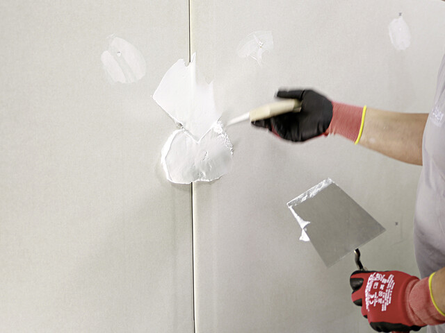 Nahaufnahme, wie Spachtelmasse auf den Fugenbereich von zwei Gipskartonplatten aufgezogen wird. Sichtbar sind zwei Hände mit einer Kelle und einem Spachtel in der Hand, von denen der Spachtel in Richtung der Fuge zeigt