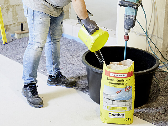 Aufnahme in der eine Person neben einem Mörtelkübel steht und Wasser in aus einem gelben Eimer in diesen Kübel schüttet. Vor dem Kübel steht eine offene Produktverpackung zum Putz- und Mauermörtel und in dem Mörtelkübel steht ein Rührgerät.