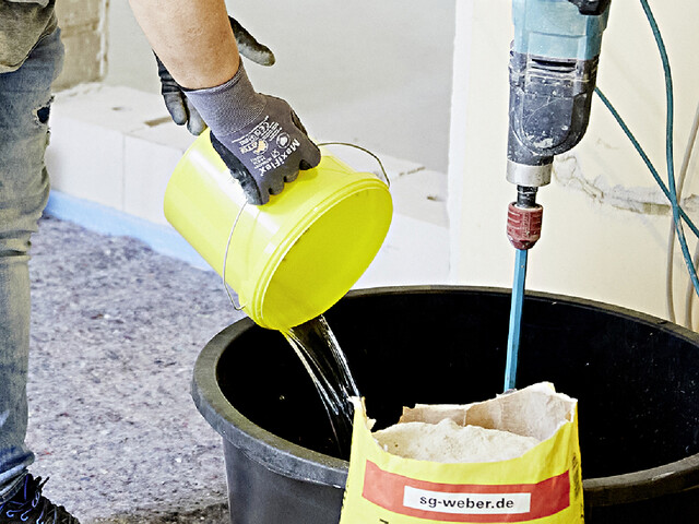 Nahaufnahme, wie eine Person Wasser aus einem gelben Eimer in einen Mörtelkübel füllt. Vor dem Mörtelkübel ist der obere Rand eines offenen Sacks vom Putz- und Mauermörtel erkennbar und im Mörtelkübel steht ein Rührstab.