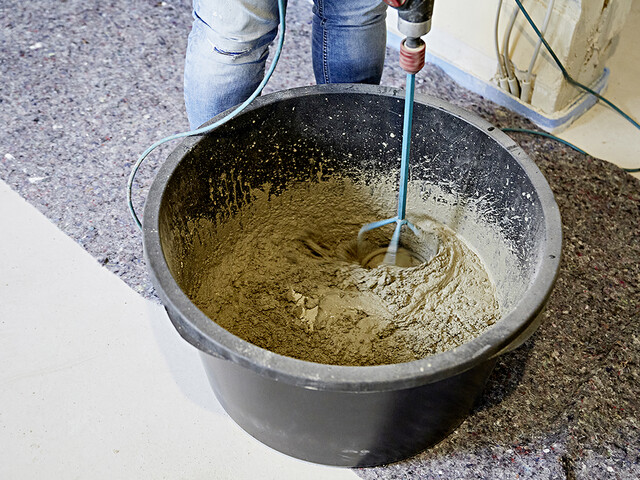 Aufnahme in einen Mörtelkübel rein, in dem der Putz- und Mauermörtel gemischt wird. Neben dem Kübel steht eine Person und im Kübel mischt ein Rührgerät die Masse.