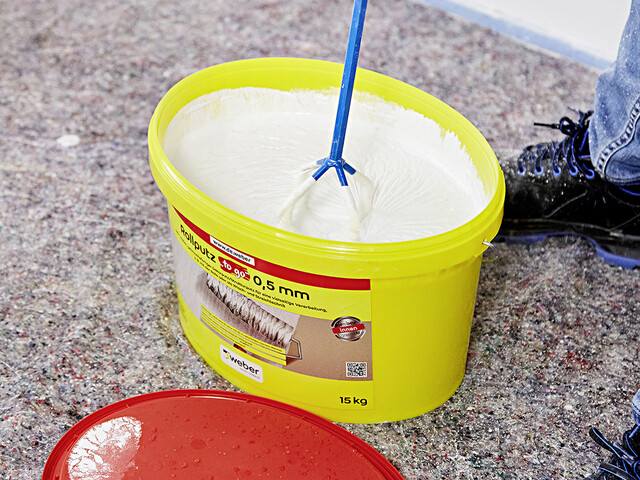 Nahaufnahme in einen gelben Produkteimer, in dem der Rollputz mit einem Rührgerät aufgerührt wird.