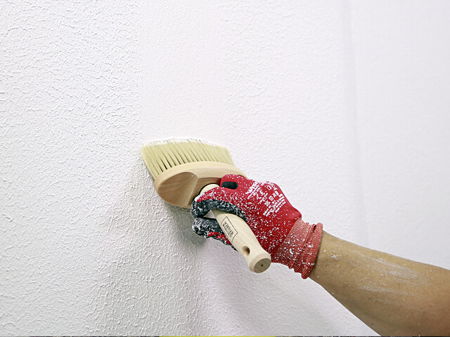 Nahaufnahme auf eine Hand, die mit einer Strukturbürste eine Wand mit Rollputz nachbearbeitet.