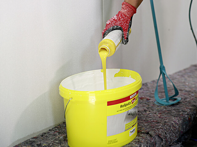 Aufnahme eines Produkteimers vom Rollputz "to go", in welchen eine Hand gelbe Farbe reinschüttet. Der Eimer steht vor einer weißen Wand und im Hintergrund ist ein Rührgerät an die Wand gelehnt.