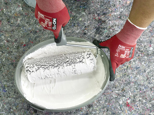 Aufnahme von oben in den Eimer mit Rollspachtel, in den eine Person eine Malerrolle eintaucht. Die Malerrolle ist zur Hälfte mit dem Rollspachtel bedeckt
