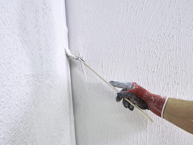 Nahaufnahme einer Innenwandecke, in welcher eine Hand mit einem Flachpinsel den Rollspachtel aufträgt