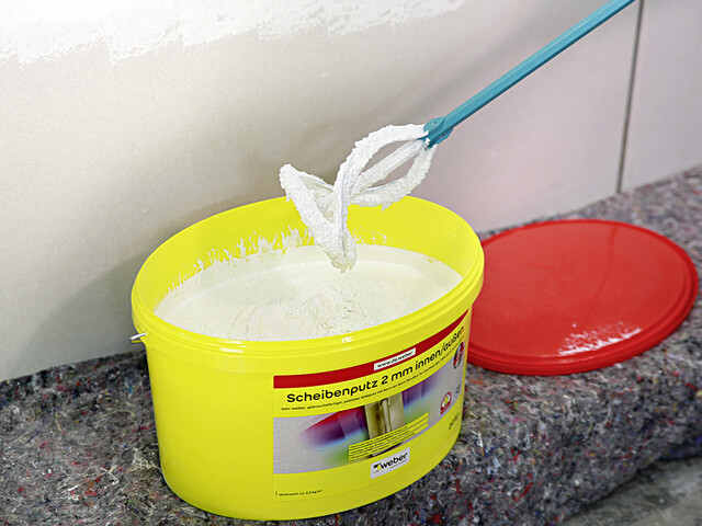 Aufnahme eines geöffneten Produkteimers vom Scheibenputz 2 mm innen/außen. In dem Eimer ist weißer Scheibenputz sichtbar und über dem Eimer wird ein Rührstab über den Putz gehalten, dessen Spitze auch voller Putz ist. Im Hintergrund hinter dem Eimer liegt der rote Deckel.