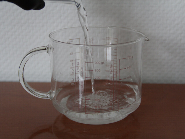 Aufnahme, wie Wasser aus einem im Bild nicht sichtbaren Gefäß in einen durchsichtigen Messbecher gegossen wird. Der Messbecher steht auf einem Holztisch vor einer weißen Wand