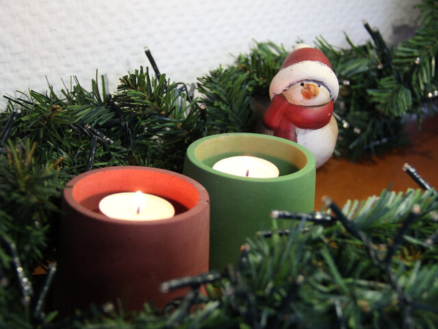 Aufnahme von Kerzenhaltern aus gefärbtem Bastelbeton in rot und grün mit Teelichtern drin. Die Kerzenhalter stehen auf einem Holztisch. Um diese herum liegt eine Lichterkette mit unechter Tannenzweig-Optik und hinter den Kerzenhaltern steht ein kleiner Schneemann aus Porzellan.