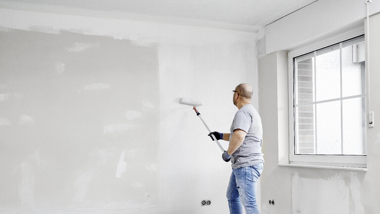 Mann steht mit einer Malerrolle vor einer Wand und trägt eine Grundierung auf die Fläche auf