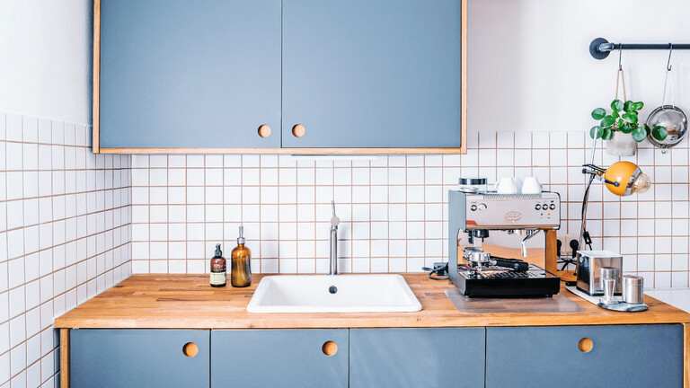 Bild von einer Küchenzeile mit weißem Fliesenspiegel. Die Küchenzeile besteht aus drei Unterschränken mit blau-grauen Türen, auf denen eine Arbeitsplatte aus Holz liegt, und einem Hochschrank mit blau-grauen Türen. In der Mitte der Arbeitsplatte ist ein weißes Waschbecken eingelassen. Links daneben stehen zwei braune Pumpflaschen und rechts daneben steht eine Espressomaschine. Die Fugen im Fliesenspiegel sind hellbraun.