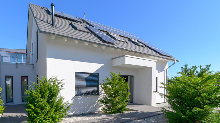 Außenansicht eines Hauses mit dunklem Dach und weißer Fassade. Davor stehen drei grüne Büsche