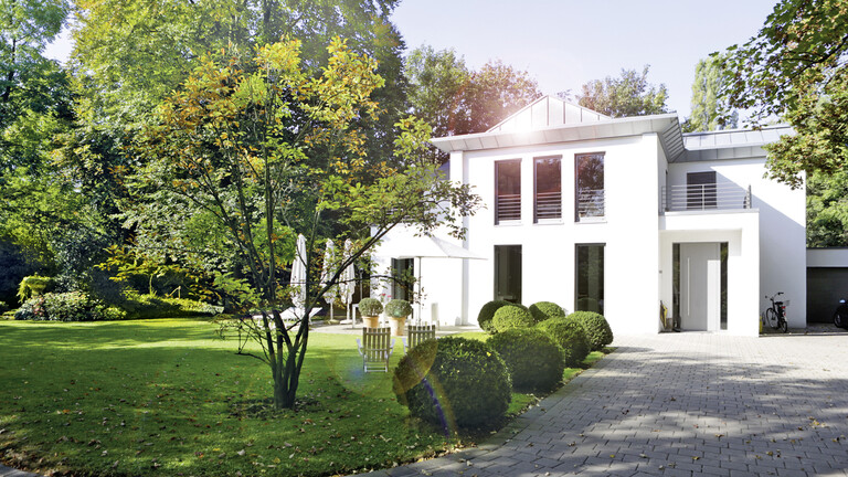 Aufnahme eines Einfamilienhauses mit weiß strahlender Fassade in einer grünen Landschaft mit Garten vor dem Haus. Im Bild sieht man einen Sonnenstrahl, der sich im Fenster des Hauses spiegelt