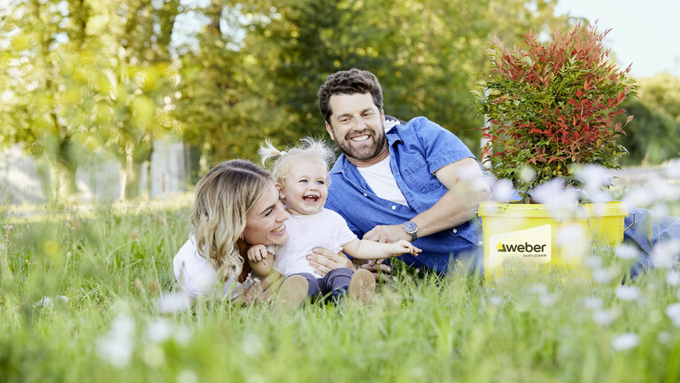 Bild von einem Mann und einer Frau, die mit einem kleinen Kind auf einer Wiese liegen. Im Hintergrund sieht man Bäume und im Vordergrund steht eine Verpackung von Saint-Gobain Weber auf dem Gras