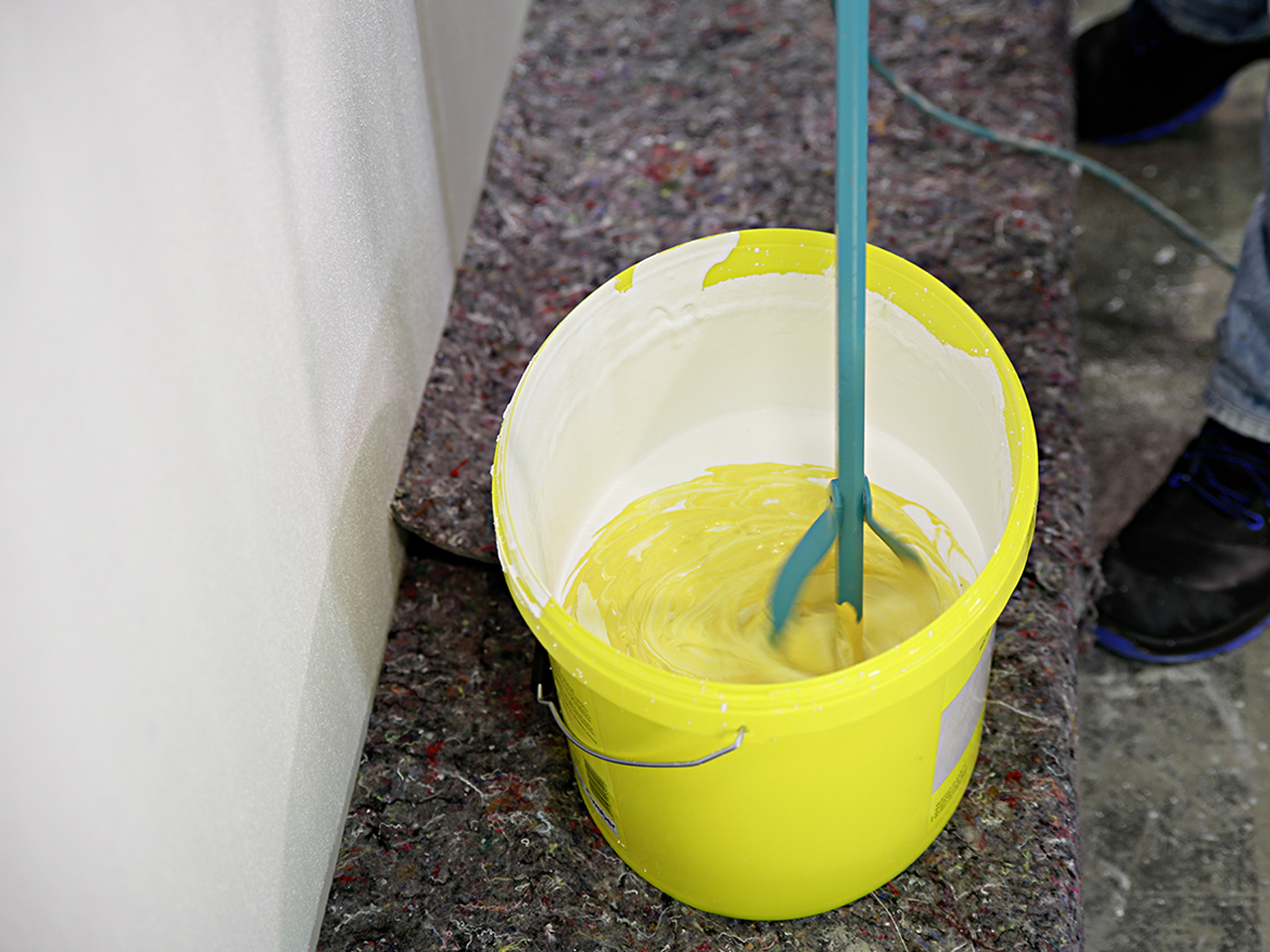 Nahaufnahme in einen gelben Produkteimer, in dem gelbe Farbe mit dem Rollputz mit einem R&uuml;hrger&auml;t vermischt wird.